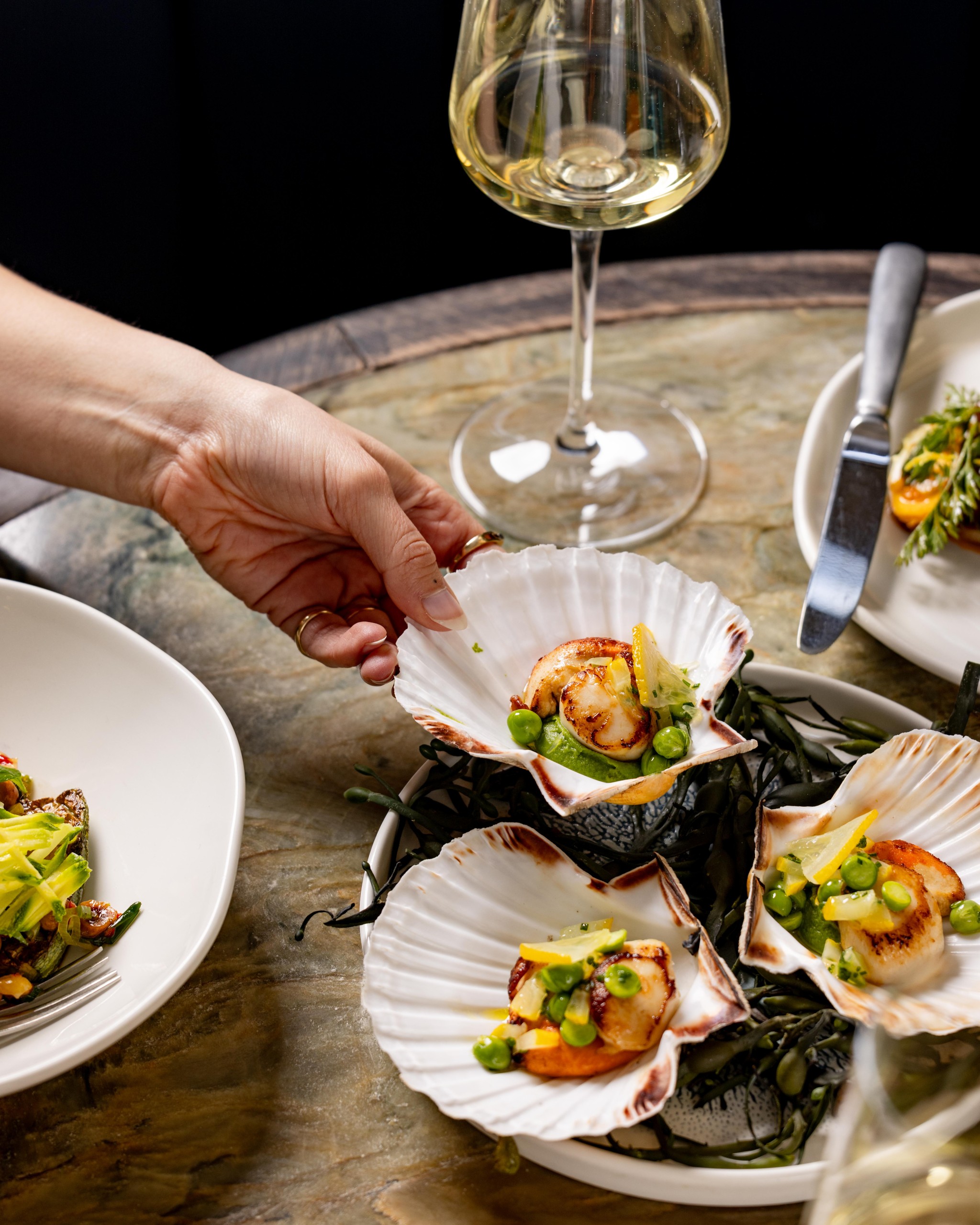A woman's hand lifting up a scallop in a shell in The Dining Room at Groucho